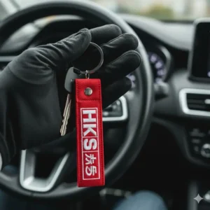 Hand holding a set of keys with a vibrant red HKS style car culture JDM keychains attached.